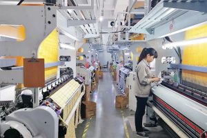 A factory worker inspecting rows of high-density weaving machines producing yellow textile labels with double-warp technology